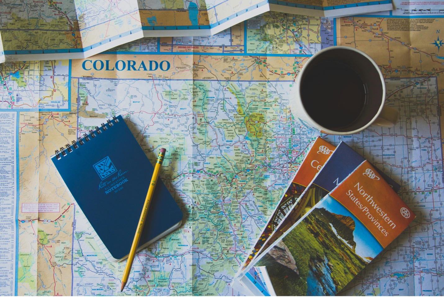 A cup of coffee notepad and folded maps all resting atop a map of Colorado while someone plots travel plans A cup of coffee notepad and folded maps all resting atop a map of Colorado while someone plots travel plans